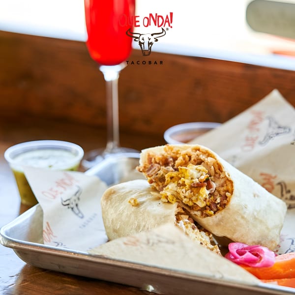 A Que Onda Tacobar burrito, cut in half, on a tray with pickled onions and a red drink.