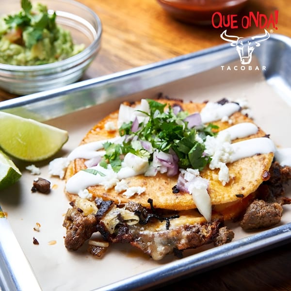 Que Onda Tacobar's carne asada taco with crema, cheese, onions, cilantro, and lime on a tray.