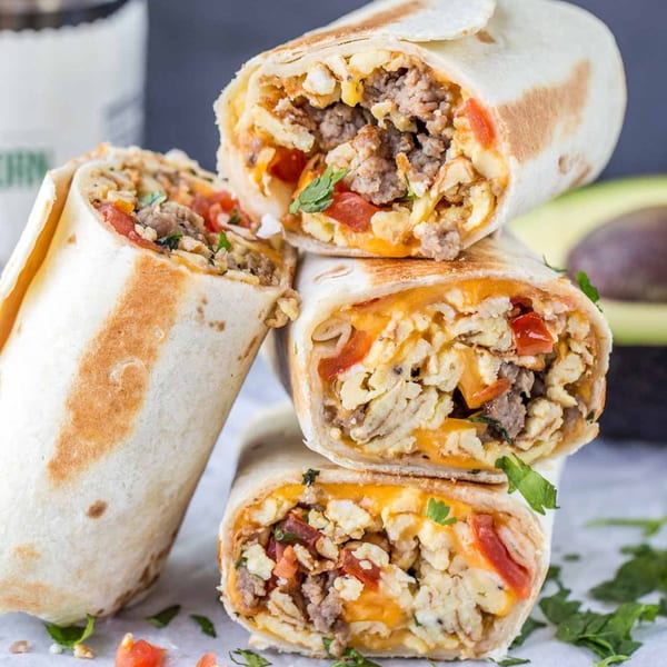 Three Que Onda Tacobar breakfast burritos, filled with eggs, sausage, cheese, and tomatoes, stacked on a table.