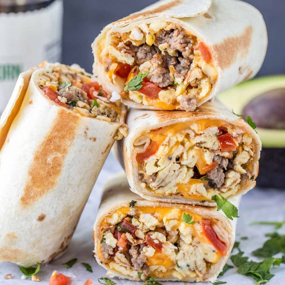 Three Que Onda Tacobar breakfast burritos, filled with eggs, sausage, cheese, and tomatoes, stacked on a table.